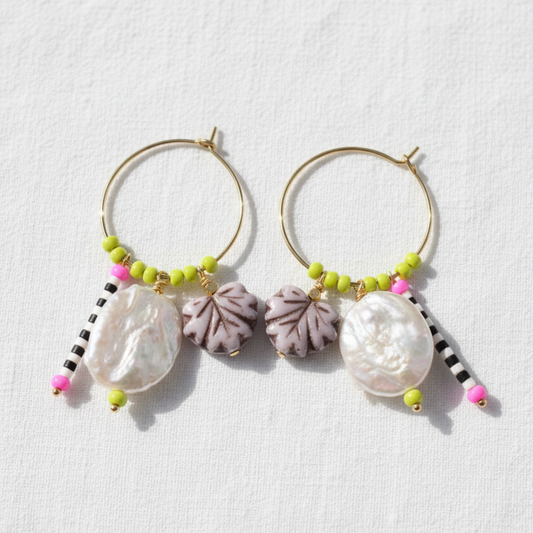 A pair of gold hoop earrings with dangling baroque shell charms, purple textured flower beads, lime green beads, and black-and-white beaded tassels, displayed on a textured white background.