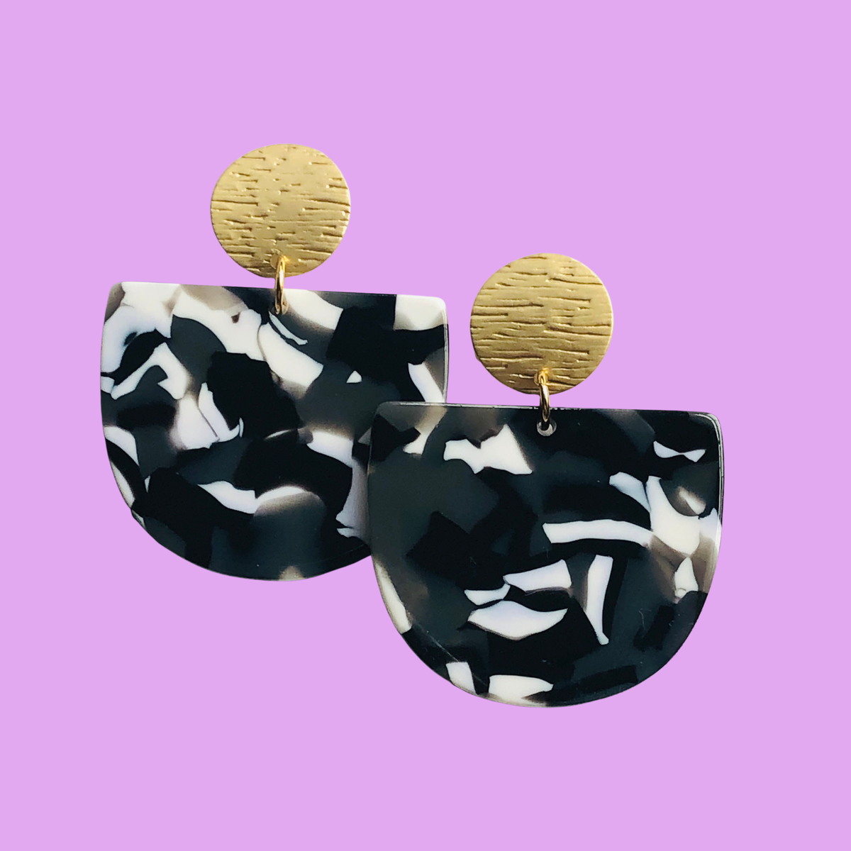 A pair of black and white swirled acrylic earrings with matte gold studs on a pale purple background.