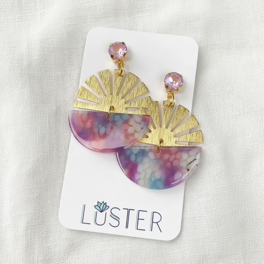 A pair of statement dangle earrings displayed on a white branded card against a soft white fabric background. Each earring has a gold-tone fan-shaped drop with a translucent marbled lower section in pink, purple, blue, and white, topped with a small faceted lavender crystal.