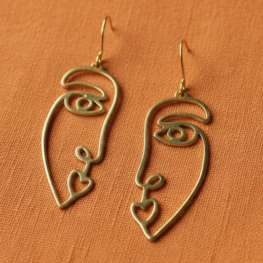 A pair of gold-toned wire dangle earrings shaped like stylized faces with eyes and heart details, displayed on a textured orange fabric background.