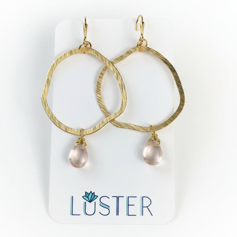 Textured brass hoop earrings with blush glass teardrop accents, displayed on a white card with the brand name 'Luster'.