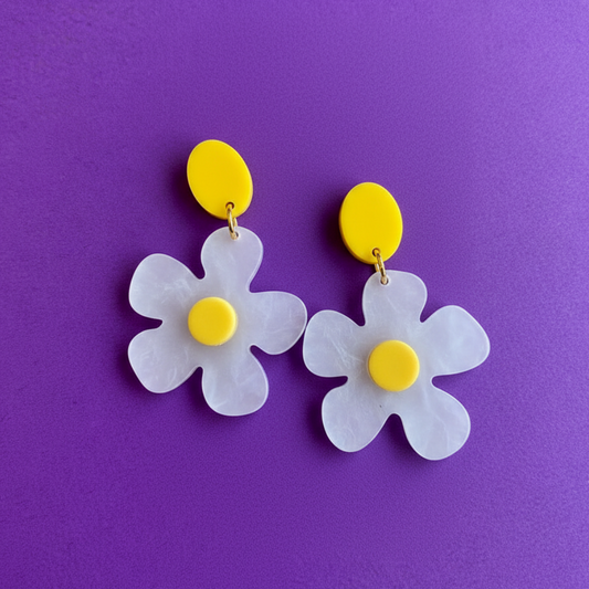 A pair of daisy-shaped drop earrings with translucent white petals, bright yellow circular studs, and small gold-tone rings, displayed on a textured purple background.