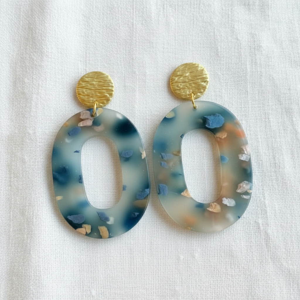 A pair of large oval drop earrings with textured gold circular studs and translucent blue, gray, and beige speckled resin dangles, displayed on a white fabric background.