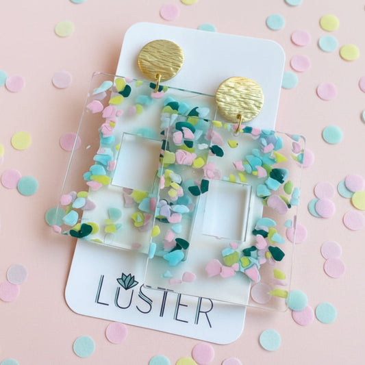 Colorful earrings on a Luster branded card with a pink background