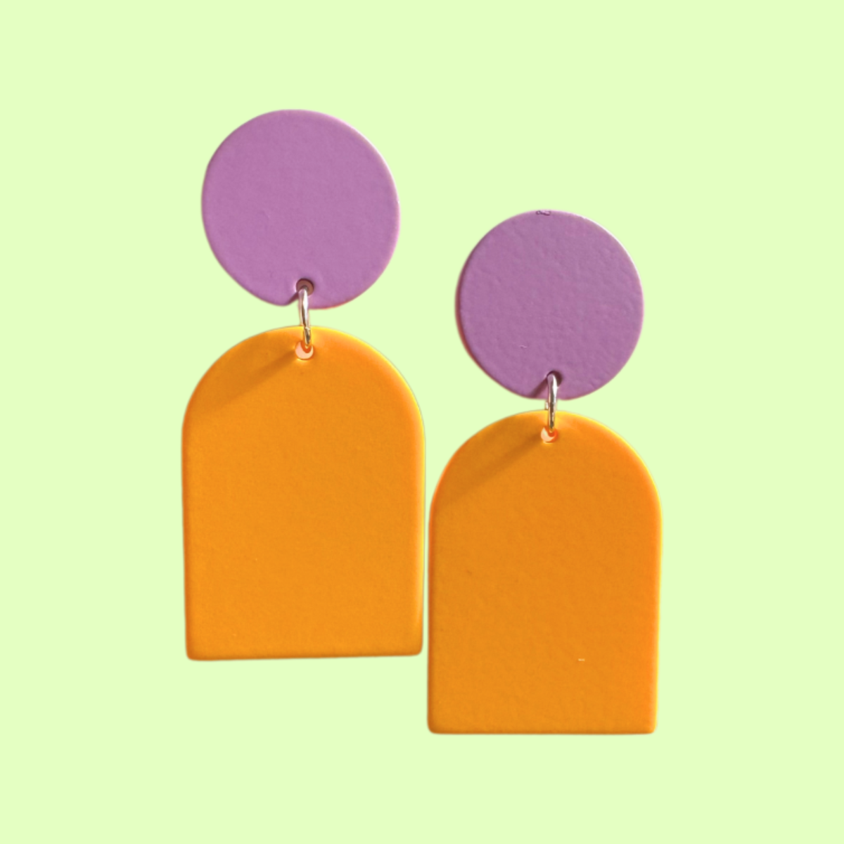 A pair of geometric drop earrings with lavender purple circular studs and bright orange arch-shaped dangles, displayed against a pale green background.