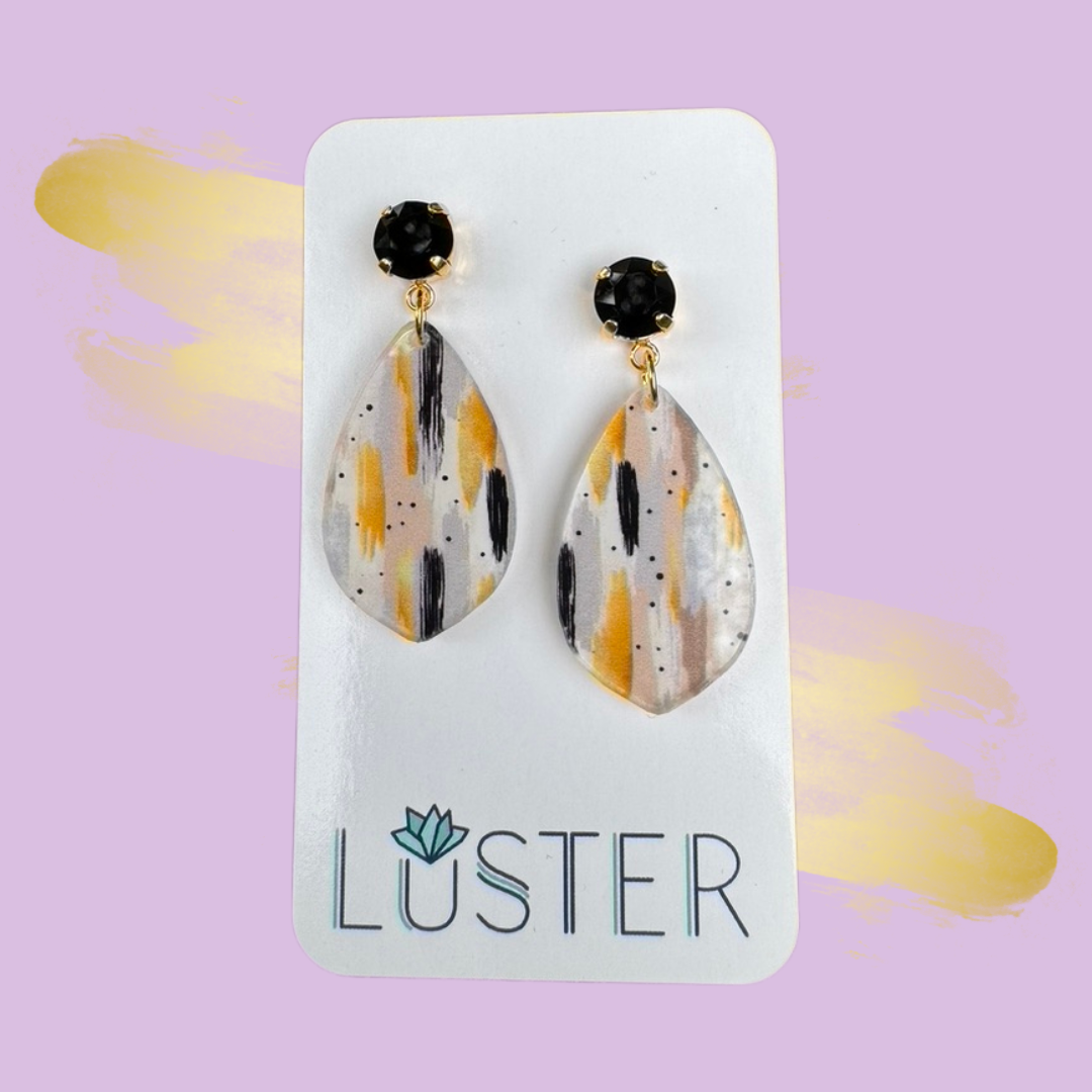 Teardrop-shaped earrings with black and gold accents on a Luster branded card against a purple background.