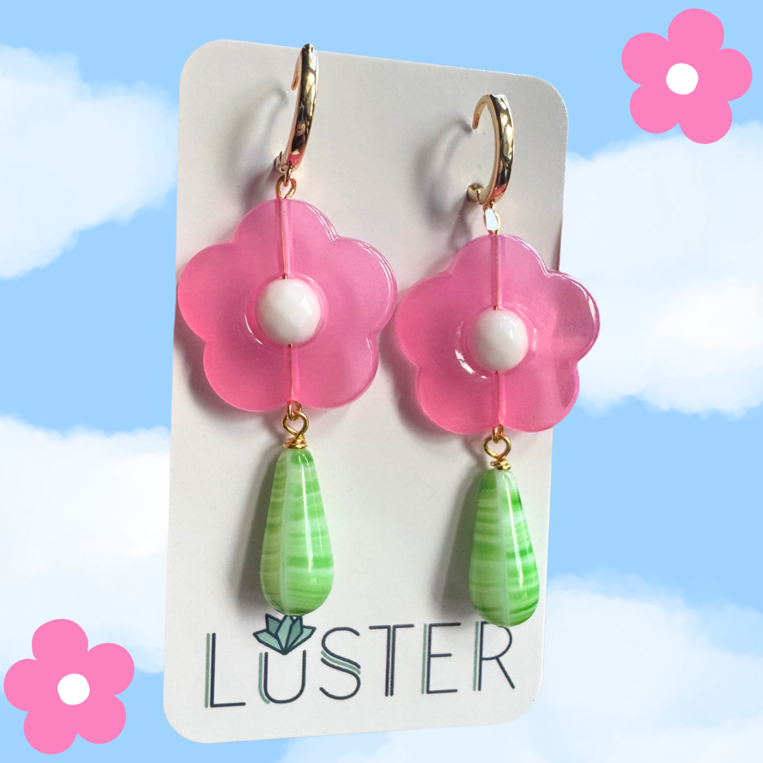 Flower-shaped earrings with pink flowers and green teardrops on a Luster branded card against a blue sky background.