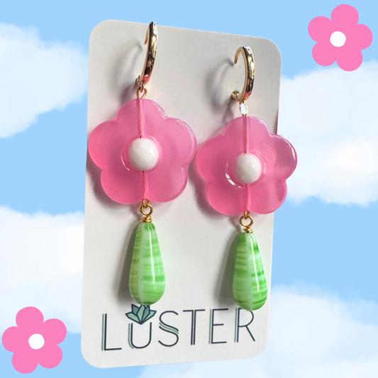 Flower-shaped earrings with pink flowers and green teardrops on a Luster branded card against a blue sky background.