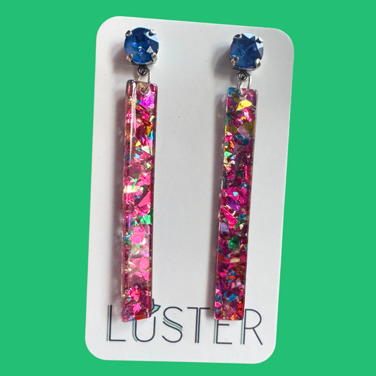 Colorful glittery earrings on a Luster branded card against a green background