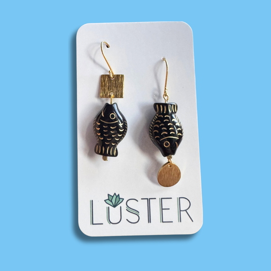 Pair of black and gold fish-shaped earrings on a Luster branded card against a blue background