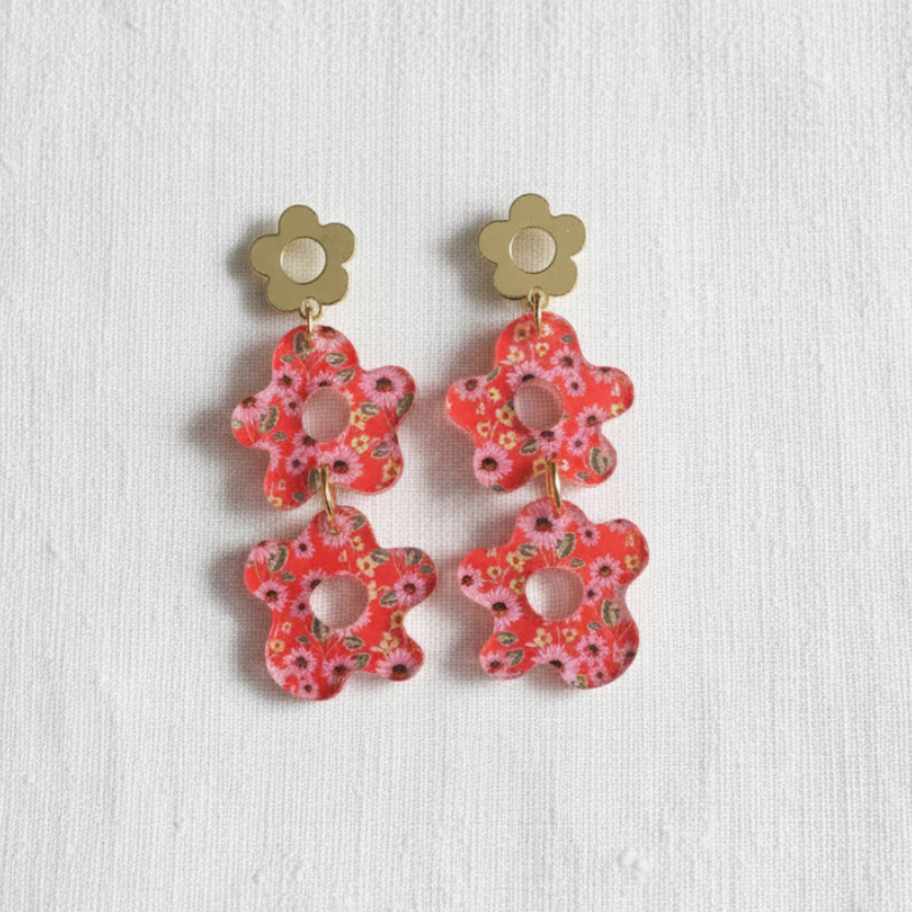 A pair of flower-shaped drop earrings with gold-tone metal studs and red-pink floral cutout pendants, displayed on a textured white fabric background.
