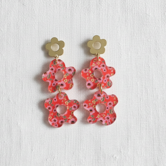 A pair of flower-shaped drop earrings with gold-tone metal studs and red-pink floral cutout pendants, displayed on a textured white fabric background.