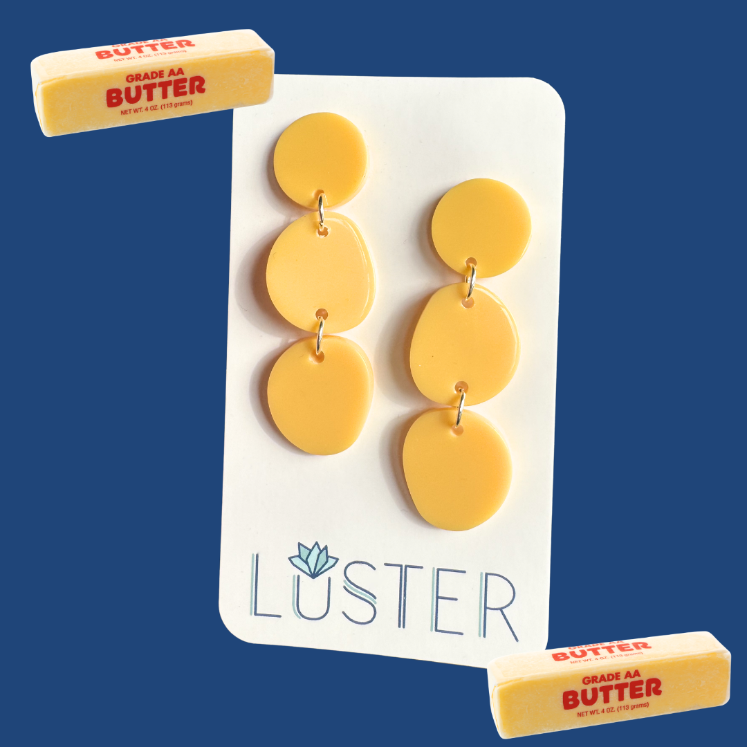 Yellow earrings on a Luster branded card with two blocks of butter on a blue background