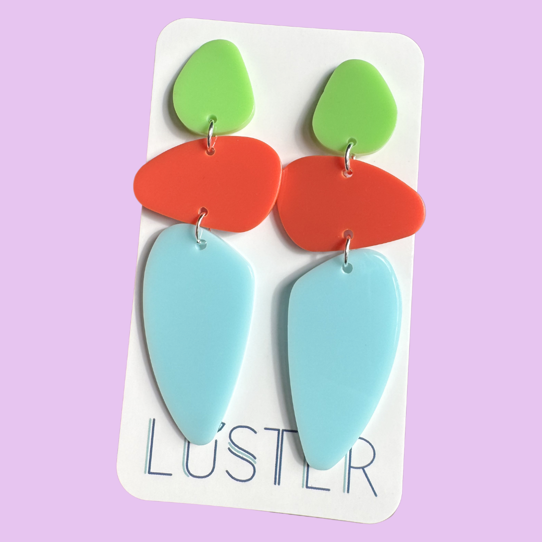A pair of colorful geometric drop earrings displayed on a white card. Each earring has three stacked shapes: a light green teardrop at the top, a coral-red rounded oval in the middle, and a large light blue teardrop at the bottom.