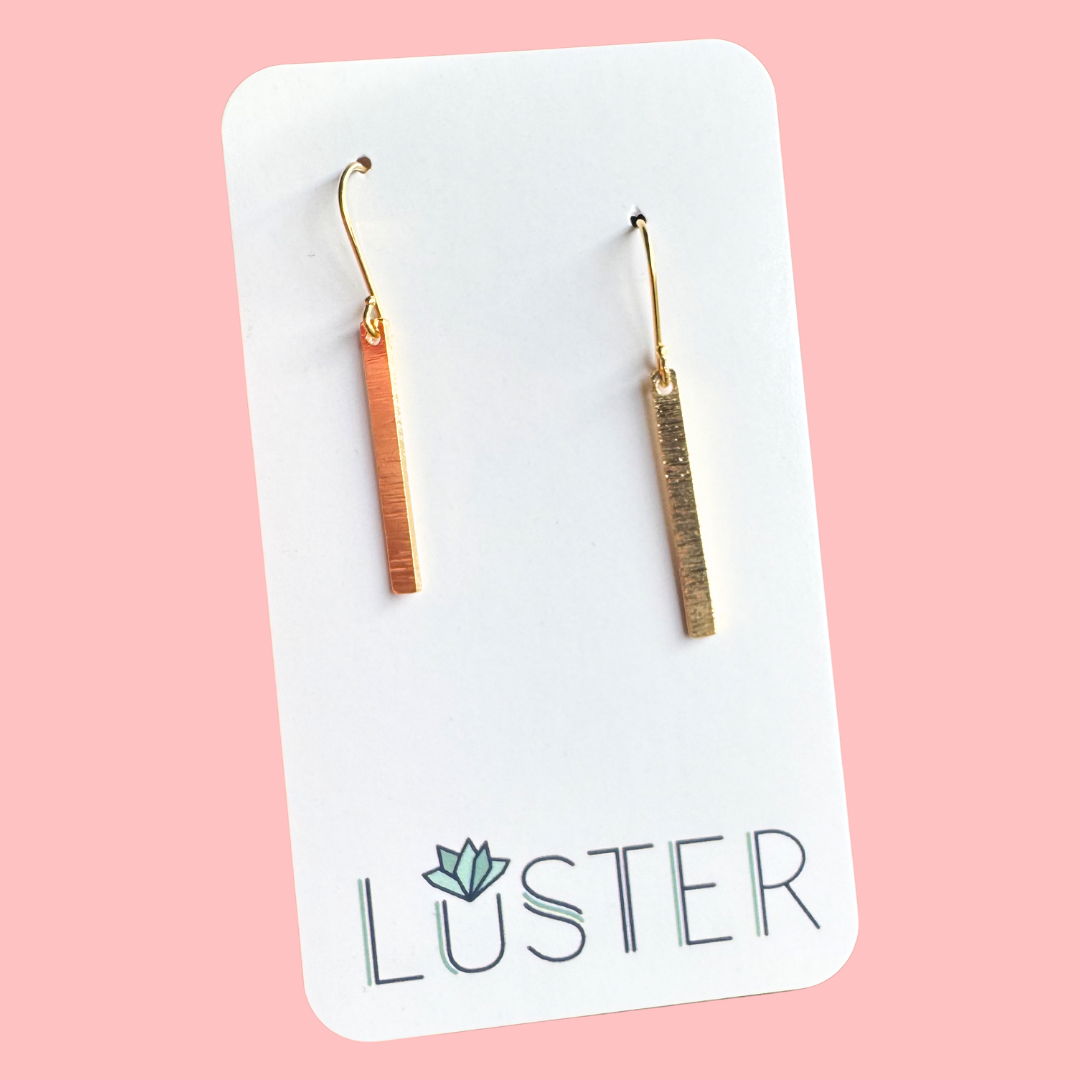 Pair of gold bar earrings on a Luster branded card against a pink background