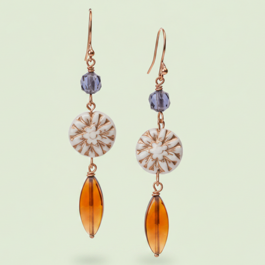 A pair of dangling earrings with rose gold hooks, small blue faceted beads, circular white and brown striped shell-like discs, and elongated amber-orange teardrop drops, shown on a pale green background.