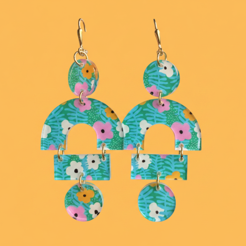 A pair of dangling earrings with a colorful floral and leaf pattern on a solid orange background. The earrings feature gold-tone leverback wires and multiple arch and circle shapes in shades of teal, green, pink, white, and yellow.