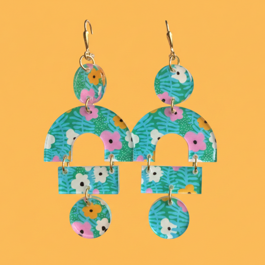 A pair of dangling earrings with a colorful floral and leaf pattern on a solid orange background. The earrings feature gold-tone leverback wires and multiple arch and circle shapes in shades of teal, green, pink, white, and yellow.
