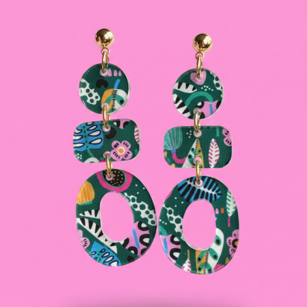 A pair of statement dangle earrings with a colorful abstract botanical pattern on a dark green background, suspended from gold-tone studs against a solid pink background.