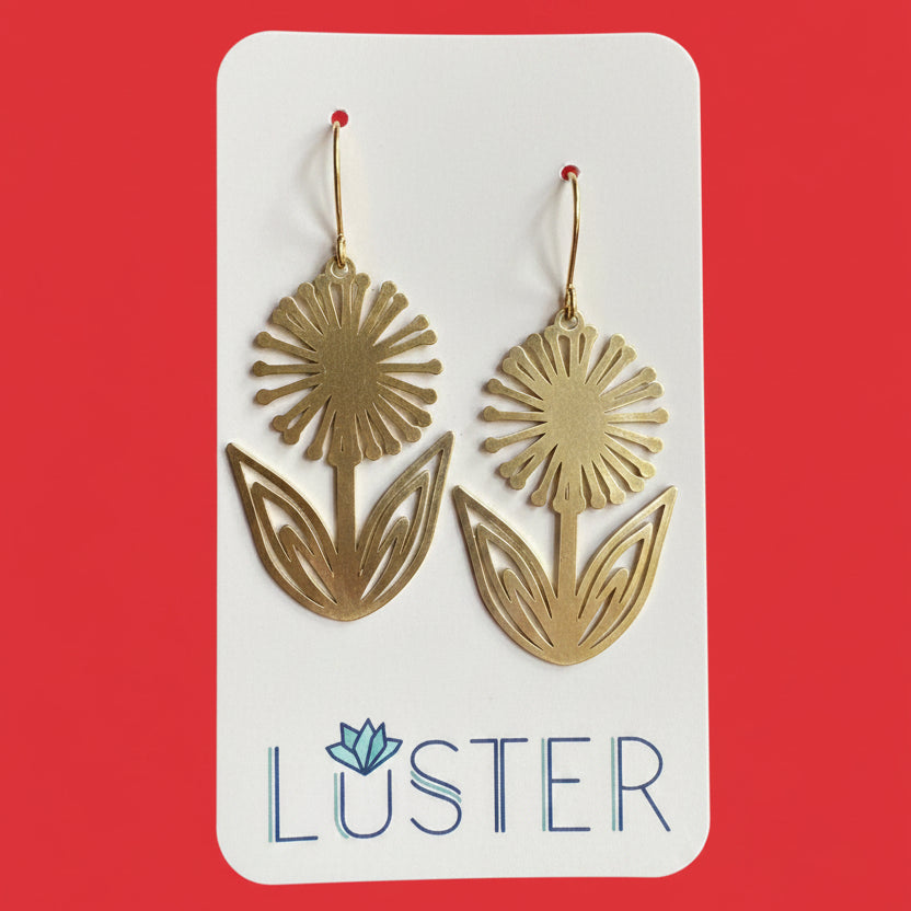 A pair of gold-tone dandelion-shaped drop earrings displayed on a white Luster brand card against a solid red background.