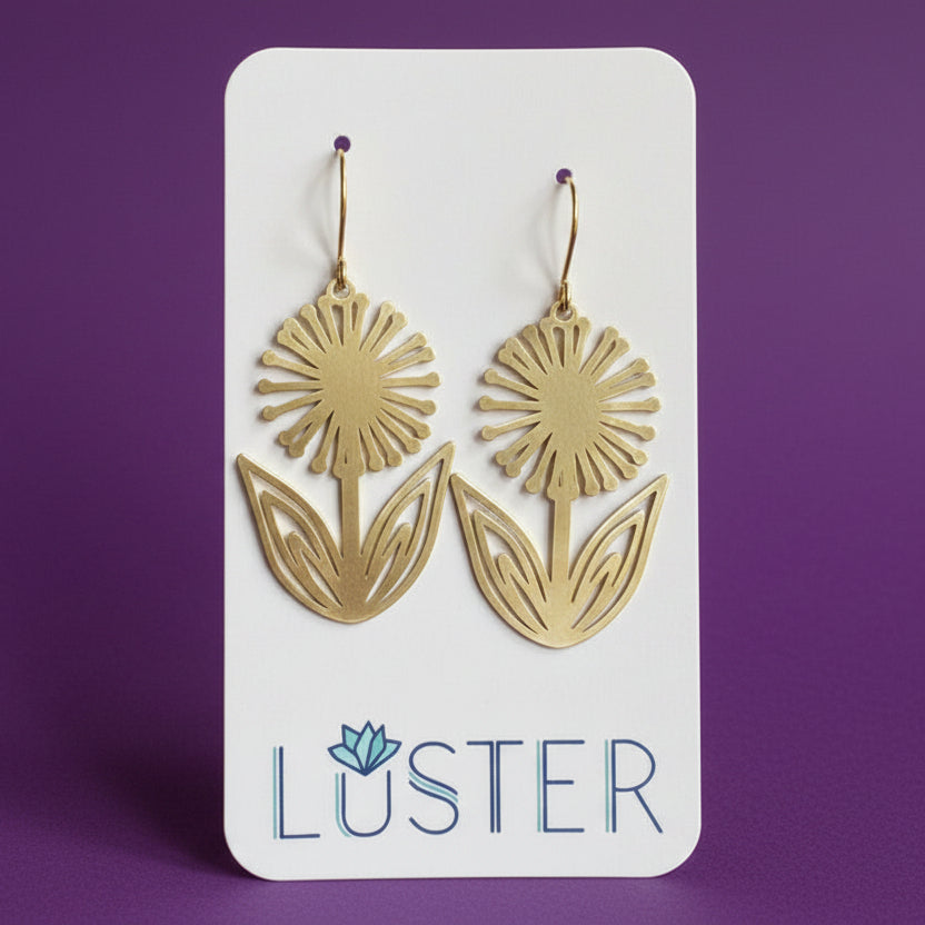 A pair of gold-tone dandelion-shaped drop earrings displayed on a white Luster brand card against a solid purple background.