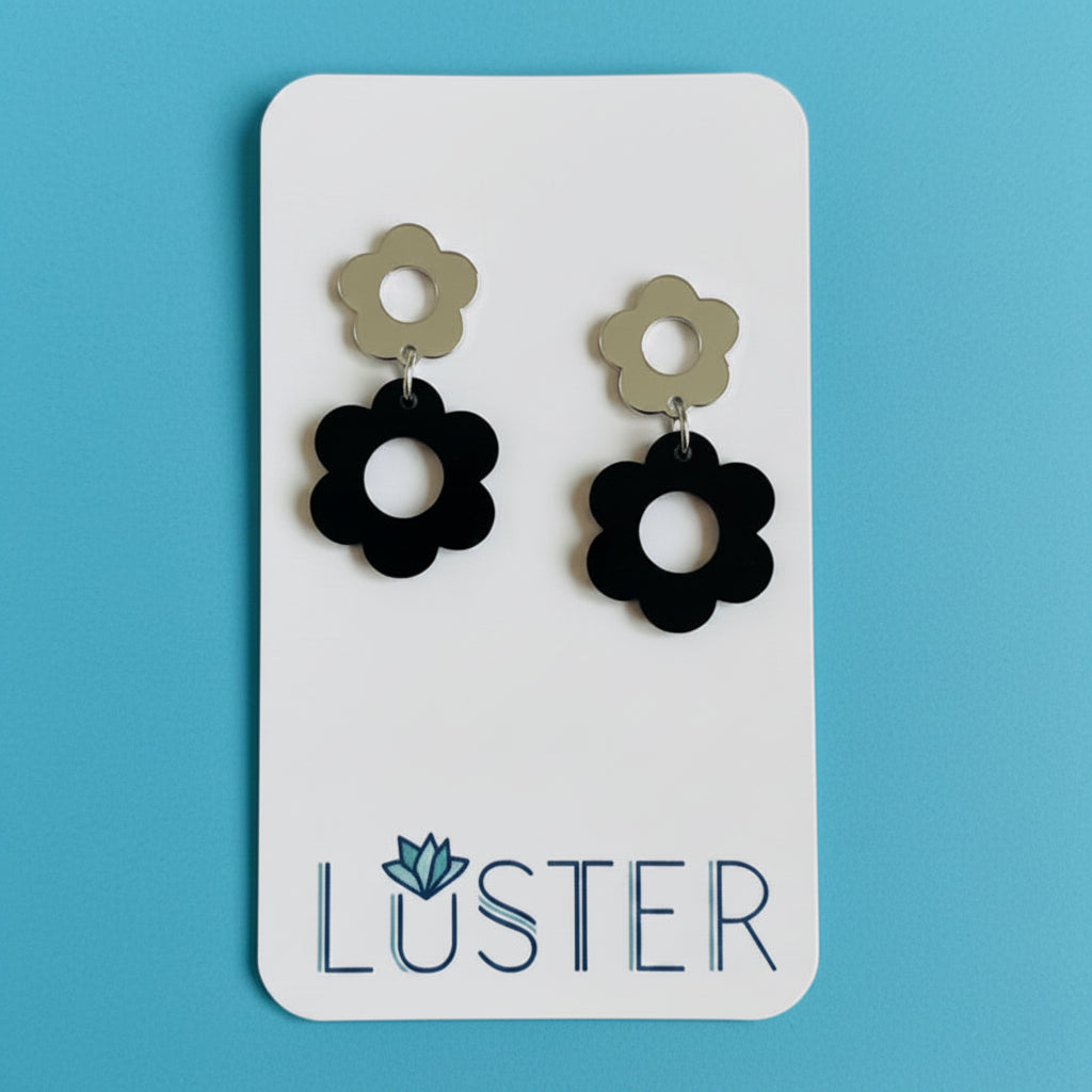 A pair of flower-shaped drop earrings displayed on a white Luster brand card against a bright blue background. Each earring has a small silver flower stud above a larger black flower.