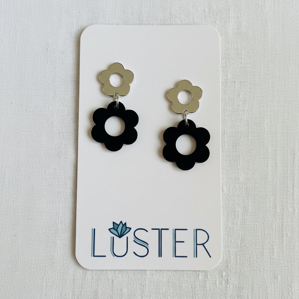 A pair of daisy earrings with black flower-shaped drops and silver flower-shaped studs, displayed on a white Luster brand card against a light textured background.