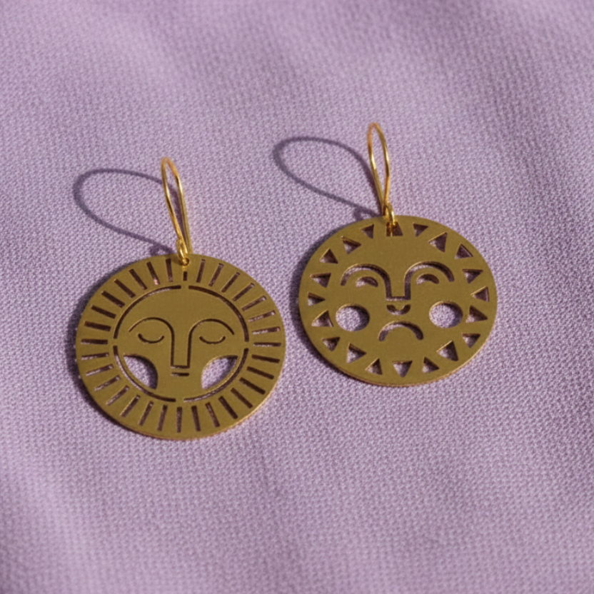 A pair of gold-toned circular drop earrings with cutout facial designs, displayed on a textured lavender fabric background.