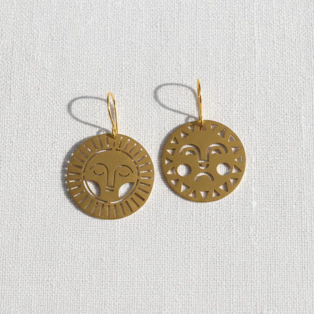 A pair of gold-toned circular dangle earrings with cutout facial details, displayed on a textured off-white fabric background.