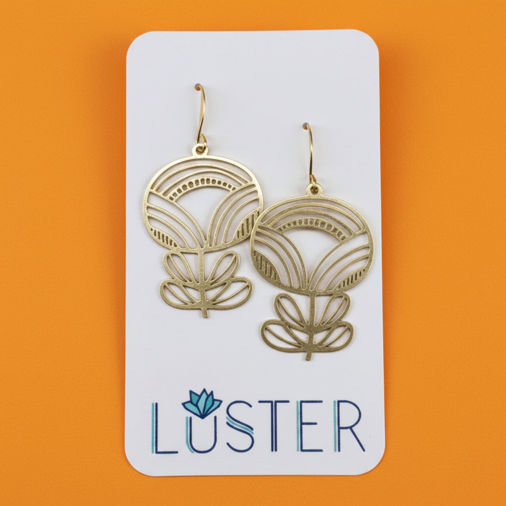 A pair of gold-toned dangle earrings with an art deco flower design, displayed on a white Luster brand card against a bright orange background.