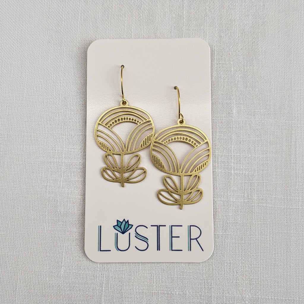 A pair of gold-toned dangle earrings with an art deco flower design, displayed on a white Luster brand card against a light gray textured fabric background.