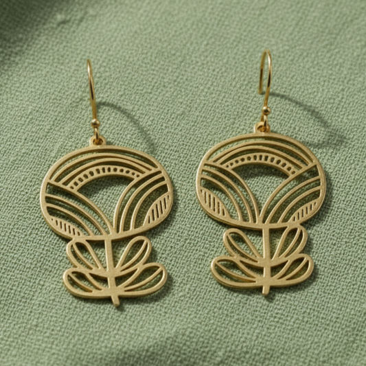 A pair of gold-toned dangle earrings with an intricate cutout floral or leaf-like design, displayed on a textured sage green fabric background.