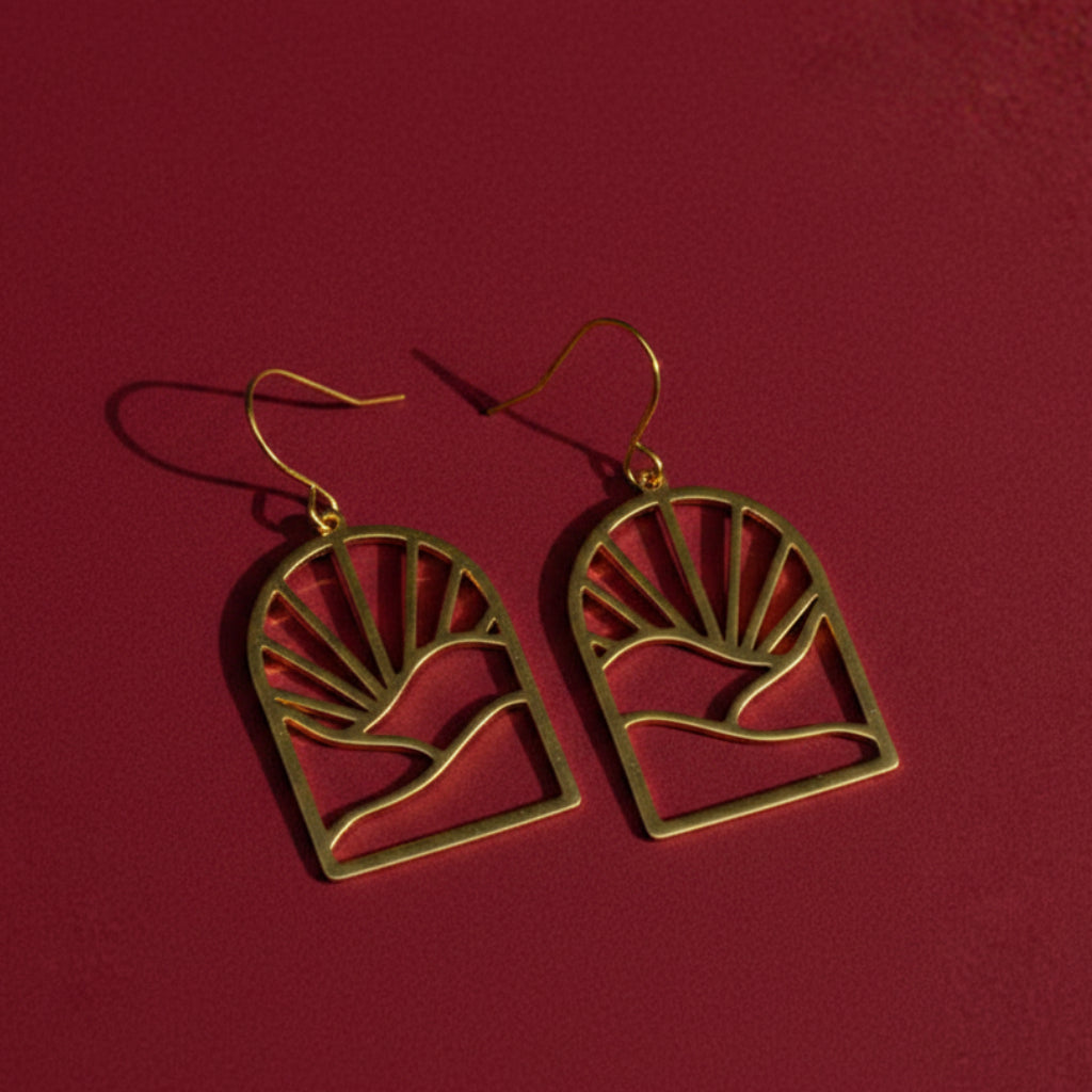 A pair of gold drop earrings with a sunburst and landscape silhouette design against a dark red background.