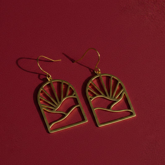 A pair of gold drop earrings with a sunburst and landscape silhouette design against a dark red background.