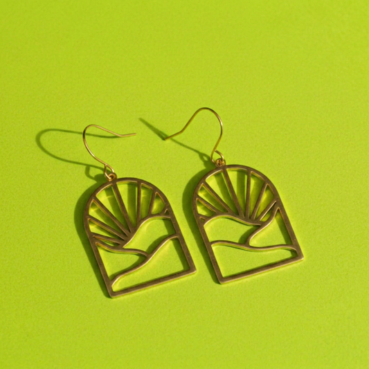 A pair of gold-tone dangle earrings with a minimalist arch design featuring a sunburst and mountain motif, displayed on a bright lime green background.