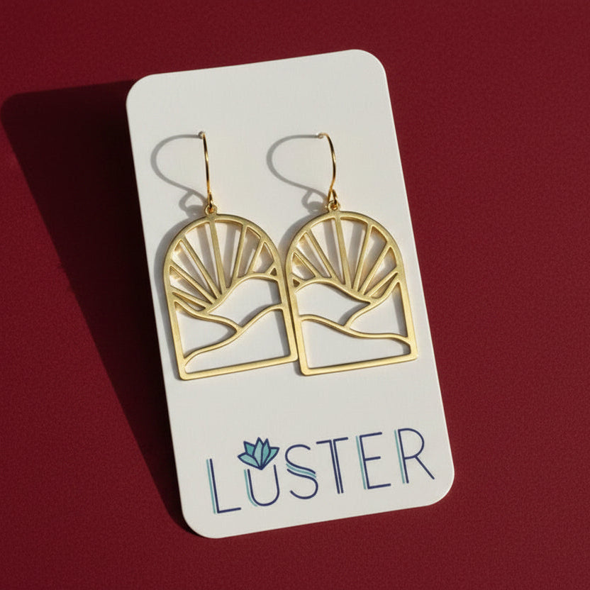 A pair of gold geometric dangle earrings displayed on a white Luster brand card against a dark red background.