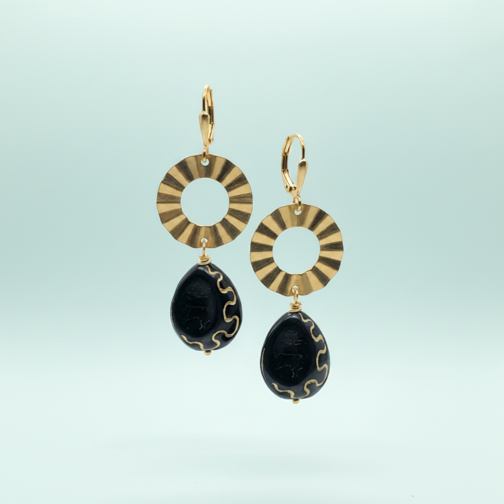 A pair of gold and black drop earrings featuring circular sunburst-patterned discs and teardrop-shaped black beads with gold wavy accents, set against a light blue gradient background.