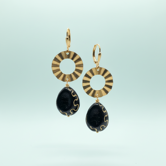 A pair of gold and black drop earrings featuring circular sunburst-patterned discs and teardrop-shaped black beads with gold wavy accents, set against a light blue gradient background.