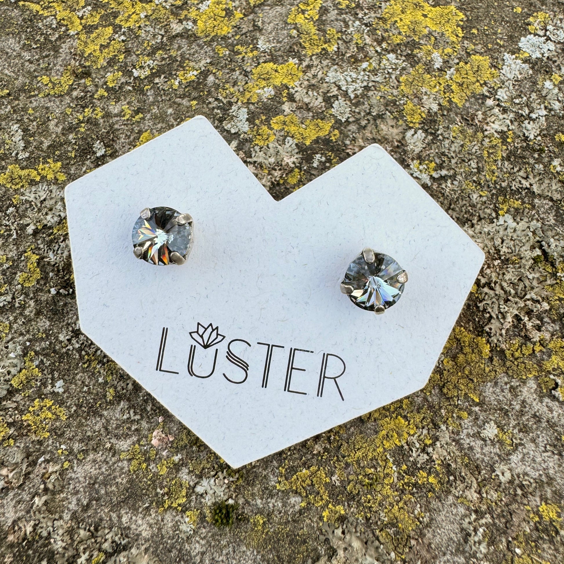 Pair of gray and silver stud earrings on a 'Luster' branded card against a textured stone background.