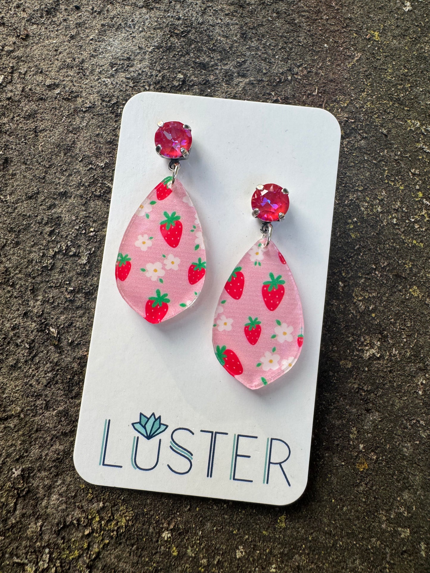Pair of pink strawberry patterned earrings on a Luster branded card against a textured surface.