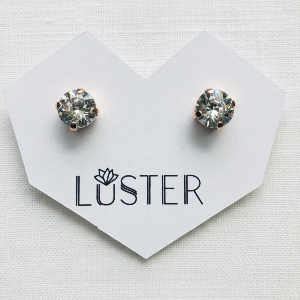 A pair of round bezel-set crystal stud earrings in a rose gold setting, displayed on a white heart-shaped card printed with the brand name 'LUSTER'.