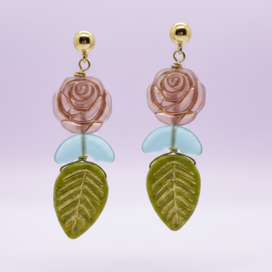A pair of dangling earrings with gold-tone posts, pink rose charms, light blue petal charms, and olive green leaf charms, shown against a soft lavender gradient background.