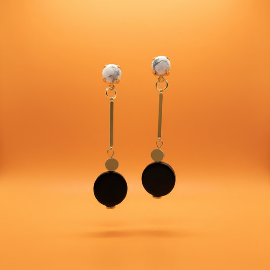 A pair of modern drop earrings with white and gray marbled studs, gold-tone metal bars, small gold discs, and large black circular pendants, set against a solid orange background.
