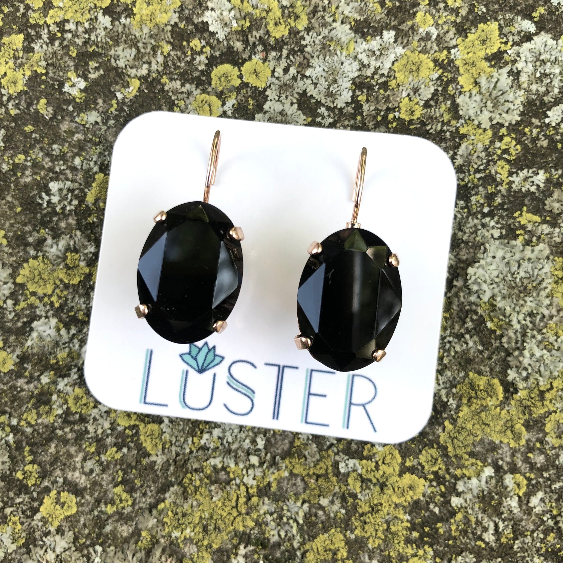 A pair of large jet black oval leverback earrings, displayed in a nickel-free finish, placed on a white card with the brand name 'LUSTER'.