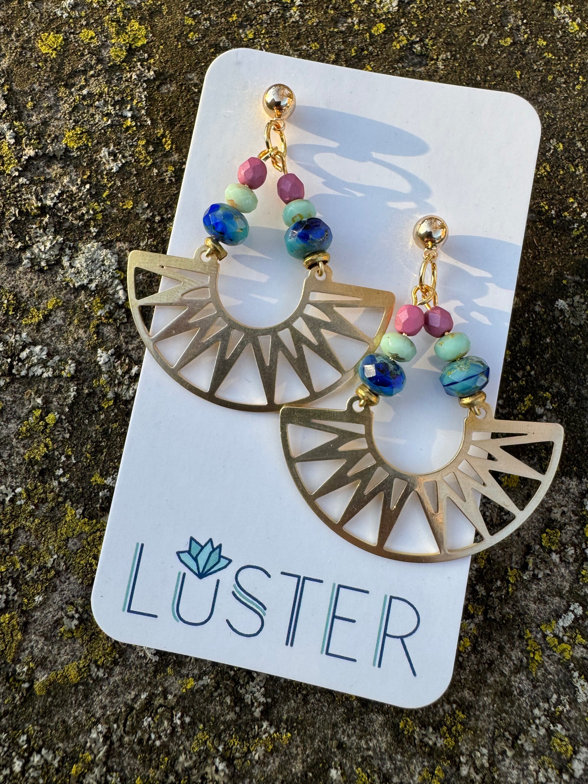 Gold earrings with colorful beads on a Luster branded card against a textured stone background