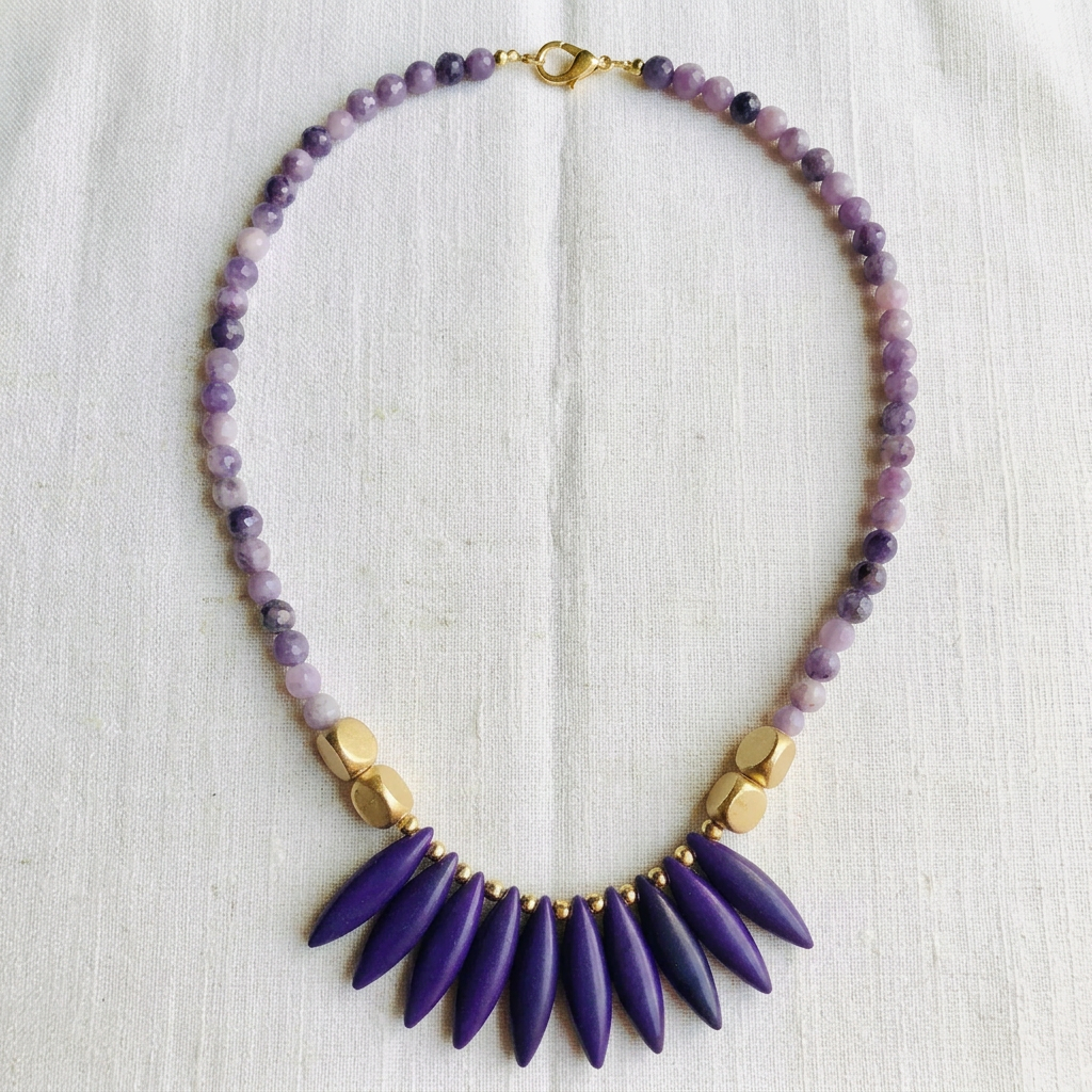 A beaded necklace laid flat on a textured off-white fabric background. The necklace features a strand of purple and lavender beads with a gold clasp, accented by gold-toned spacer beads, and a prominent fan of deep purple teardrop-shaped beads at the center.