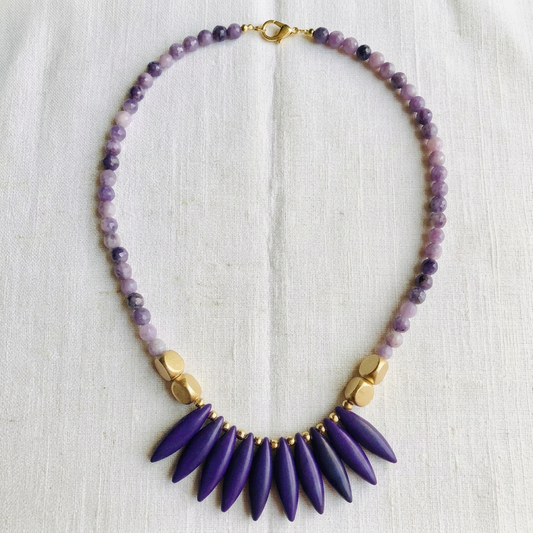 A beaded necklace laid flat on a textured off-white fabric background. The necklace features a strand of purple and lavender beads with a gold clasp, accented by gold-toned spacer beads, and a prominent fan of deep purple teardrop-shaped beads at the center.