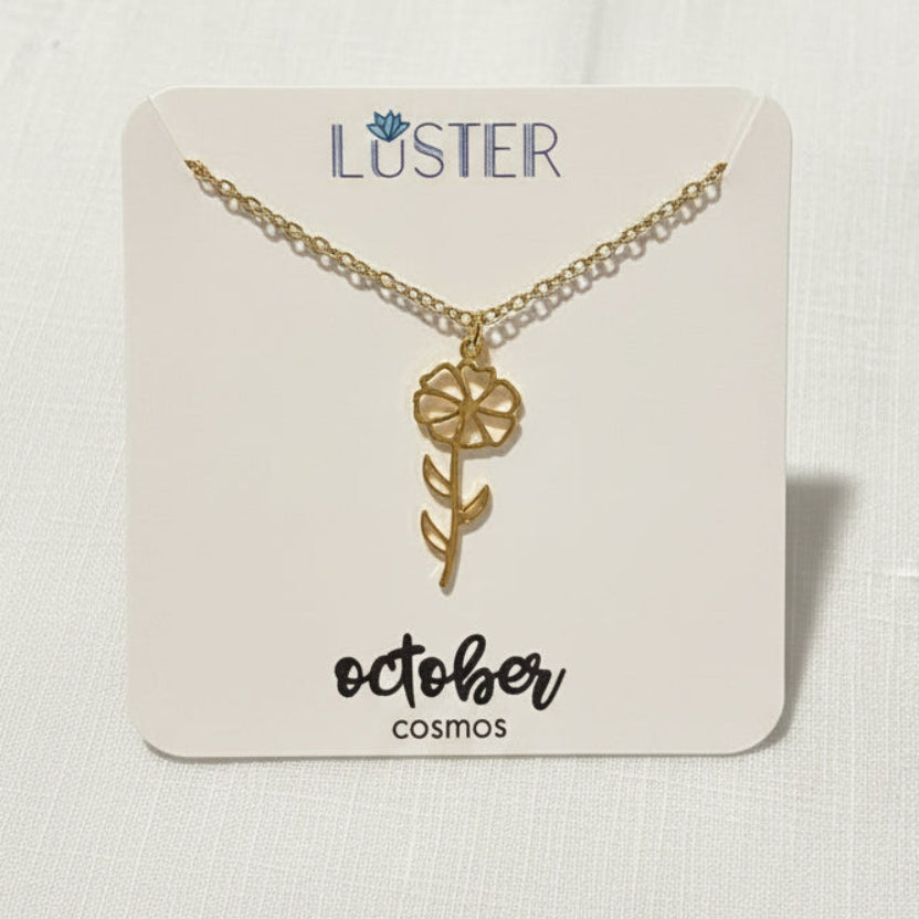 A gold flower pendant necklace on a delicate chain, displayed on a white Luster brand card with the text 'october cosmos' at the bottom, set against a light textured fabric background.