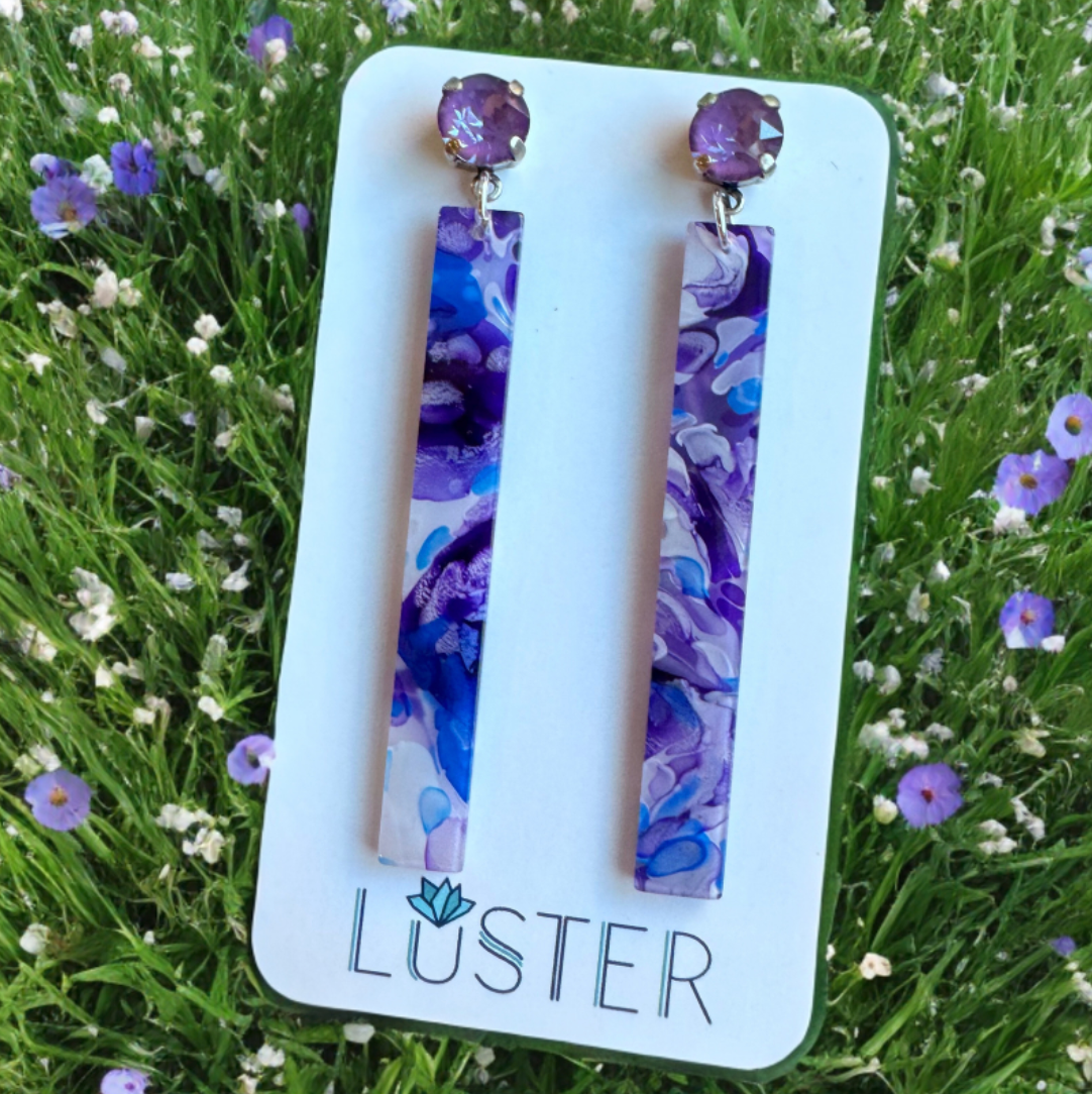 Pair of purple floral earrings on a Luster branded card with grass and flowers in the background