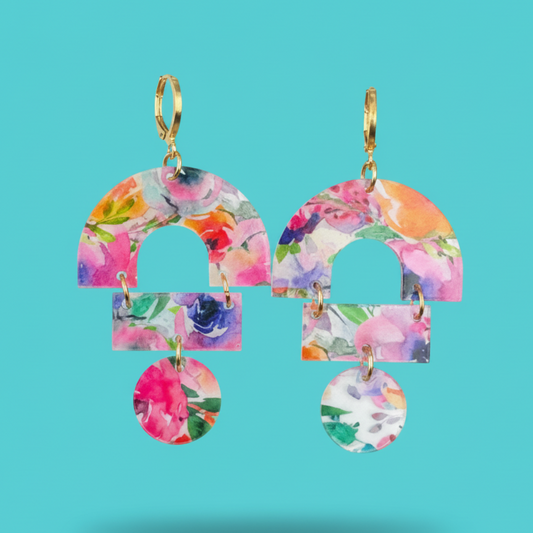 A pair of gold-tone hoop earrings with layered arch and rectangular dangles covered in a colorful floral print, shown against a solid turquoise background.
